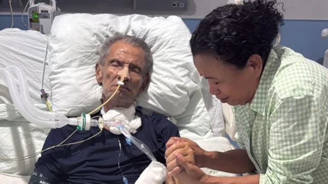 Brooklyn Rivera está acostado en una cama de hospital con un tubo conectado a su nariz. Nancy Elizabeth Henríquez, quien está parada a su lado, le agarra una mano con sus dos manos mientras cierra sus ojos